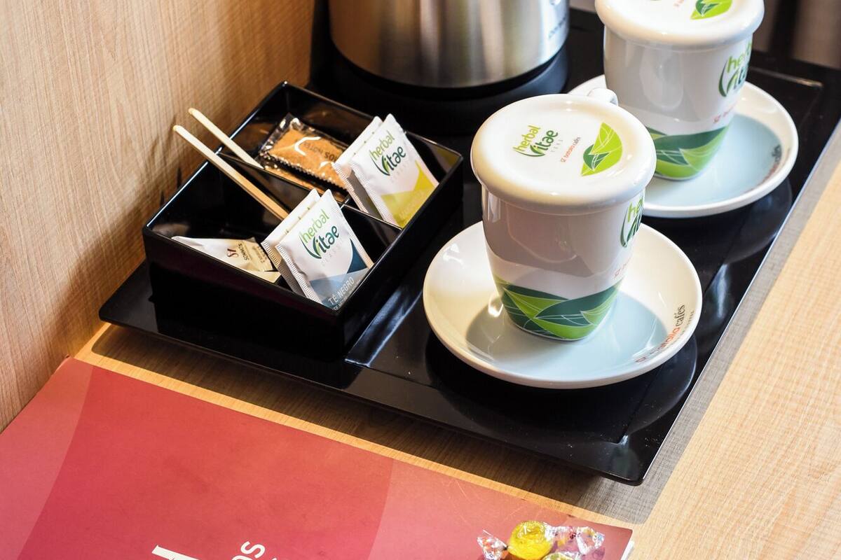Habitación de hotel con un minibar que contiene bolsitas de té de hierbas, una tetera y dos tazas con un logo de hoja verde. Un cuaderno rojo y un lápiz están sobre la mesa.