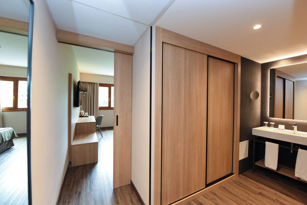 Una habitación de hotel moderna con un armario de madera, un baño con un espejo grande y una zona de escritorio con una silla.