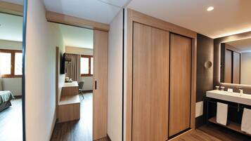 A modern hotel room with a wooden wardrobe, a bathroom with a large mirror, and a desk area with a chair.