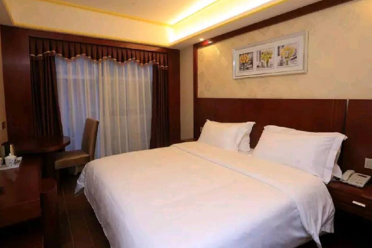 A hotel room with a large bed, a desk with a chair, a telephone, and a painting on the wall.