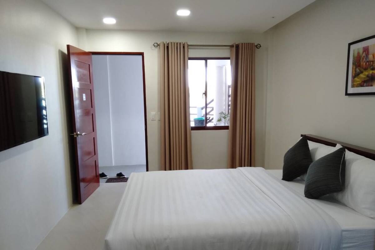 A bedroom with a bed, a TV mounted on the wall, a door, and a window with curtains.