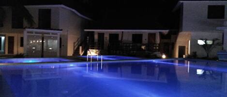 A swimming pool with a blue-lit edge, surrounded by buildings with large windows and a staircase leading to a covered area.
