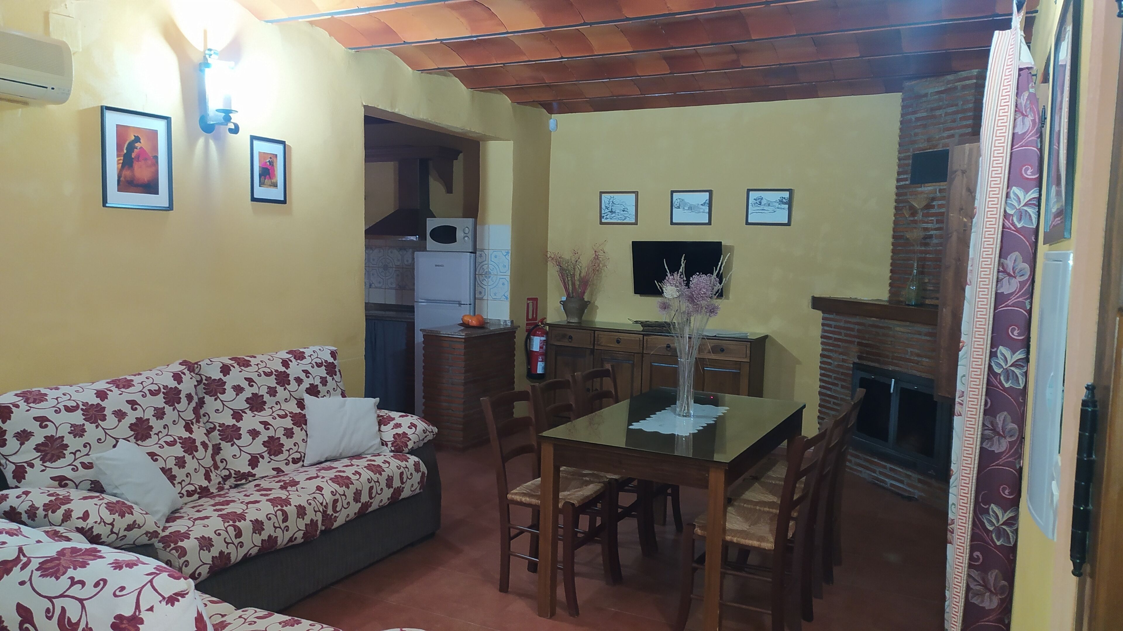 Uma sala de estar com sofá, uma mesa de jantar com cadeiras, lareira e uma cozinha com geladeira e fogão.