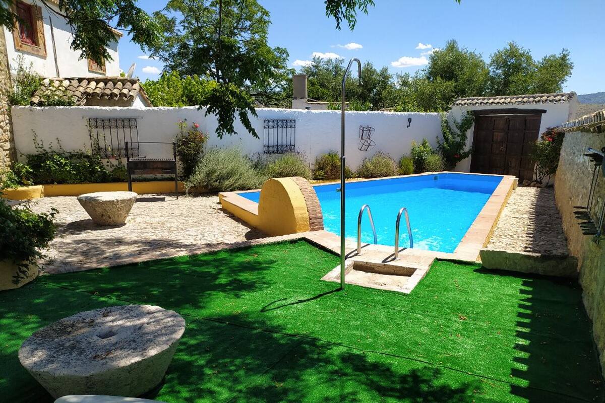 Una piscina con borde de piedra, rodeada por un muro blanco con detalles decorativos y un césped verde.