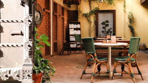 An outdoor patio with a round table and four wicker chairs, surrounded by potted plants and a brick wall. - Mangaldas Ni Haveli II by The House of MG (Ahmedabad)