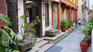 A narrow street with a building featuring a traditional entrance with a wooden door and steps leading up to it. There are potted plants along the sidewalk and a person walking in the background. - Mangaldas Ni Haveli II by The House of MG (Ahmedabad)