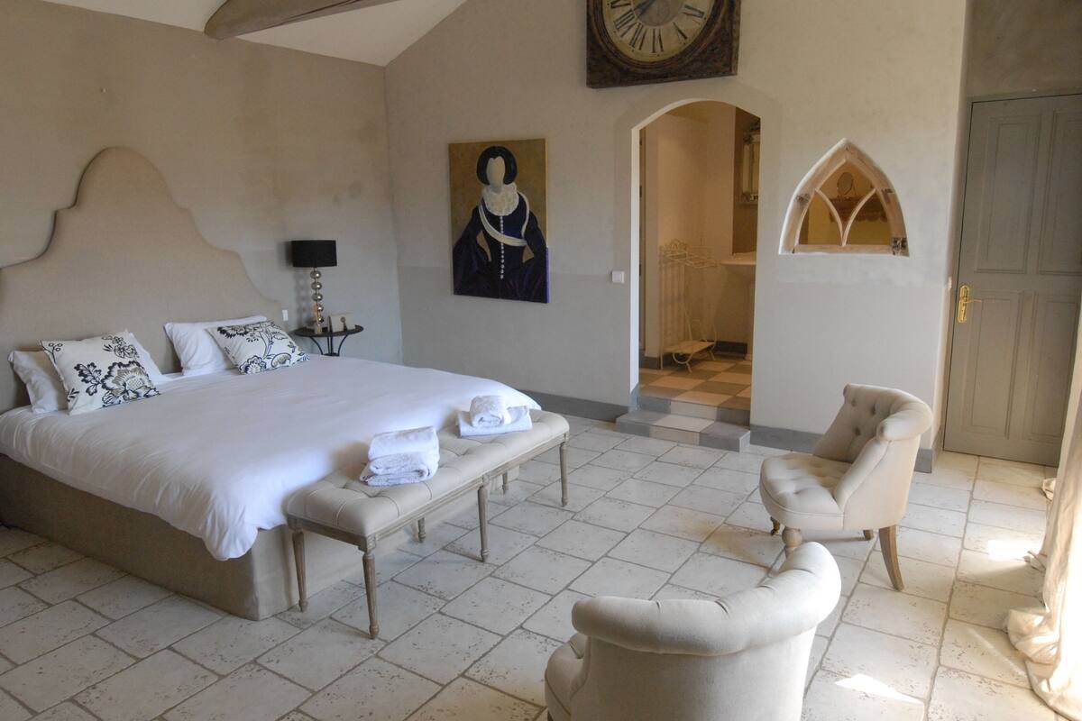 Une chambre à coucher avec un grand lit, un banc, une chaise, une horloge et un tableau.