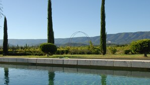 Une piscine aux eaux limpides où se reflètent les environs, bordée de haies soigneusement taillées et de grands arbres, avec une chaîne de montagnes en toile de fond.