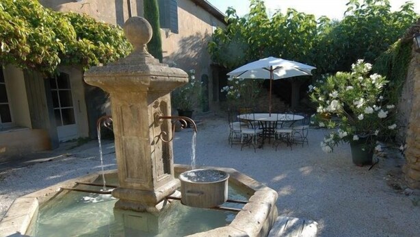 Une fontaine en pierre dont l’eau jaillit du sommet, entourée d’une zone recouverte de gravier. On y trouve une table et des chaises sous un parasol, ainsi qu’un pot de fleurs avec des fleurs blanches sur la droite.