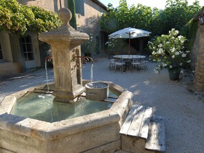 Una fontana in pietra con acqua che zampilla da una colonna centrale, circondata da un'area ghiaiosa con una panchina in legno e un tavolo con sedie.