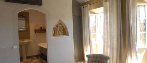 A bathroom with a checkered floor, a sink, and a mirror. There is a chair in the room and a window with curtains.