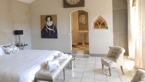 Une chambre à coucher avec un grand lit, une chaise, un banc, une horloge, un tableau, et une salle de bain visible à travers une arche.