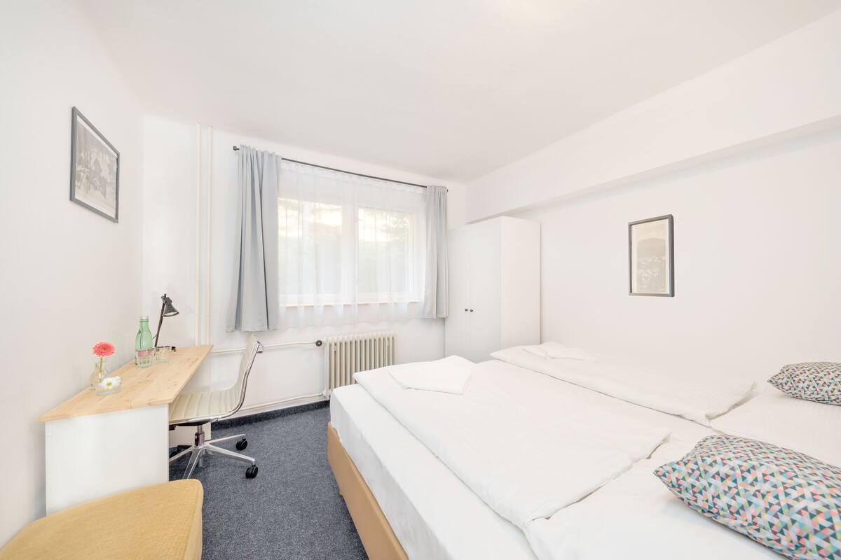 A hotel room with a bed, a desk, a chair, a radiator, and a window with curtains.