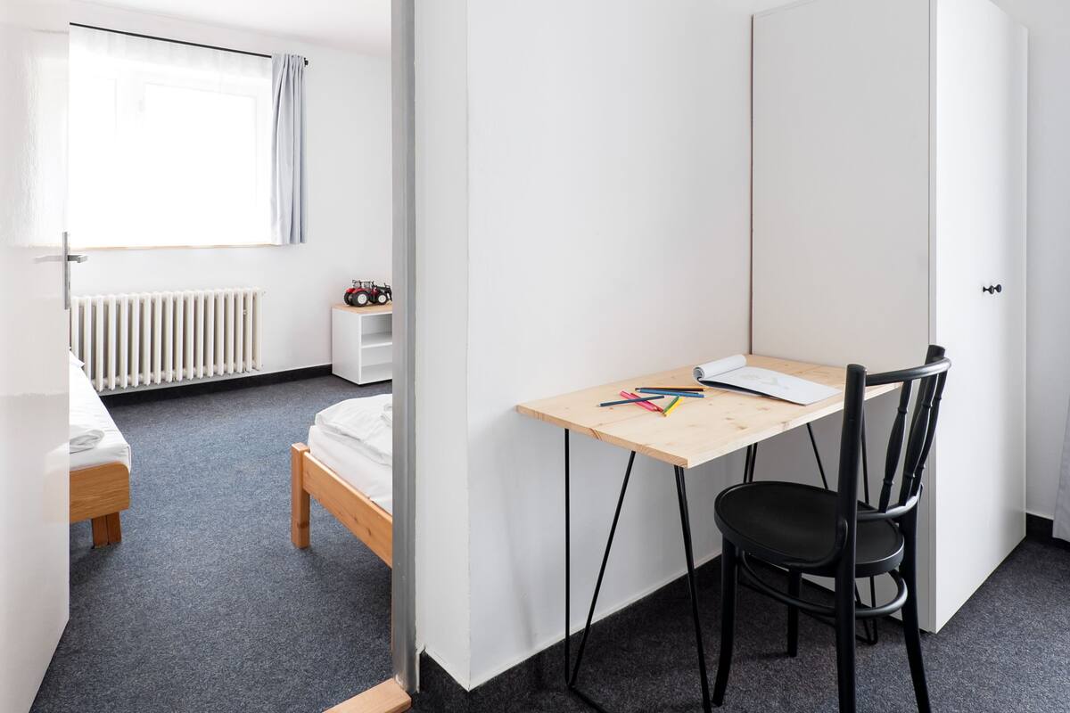 A compact room with a bed, a desk with a chair, a radiator, and a window with curtains.