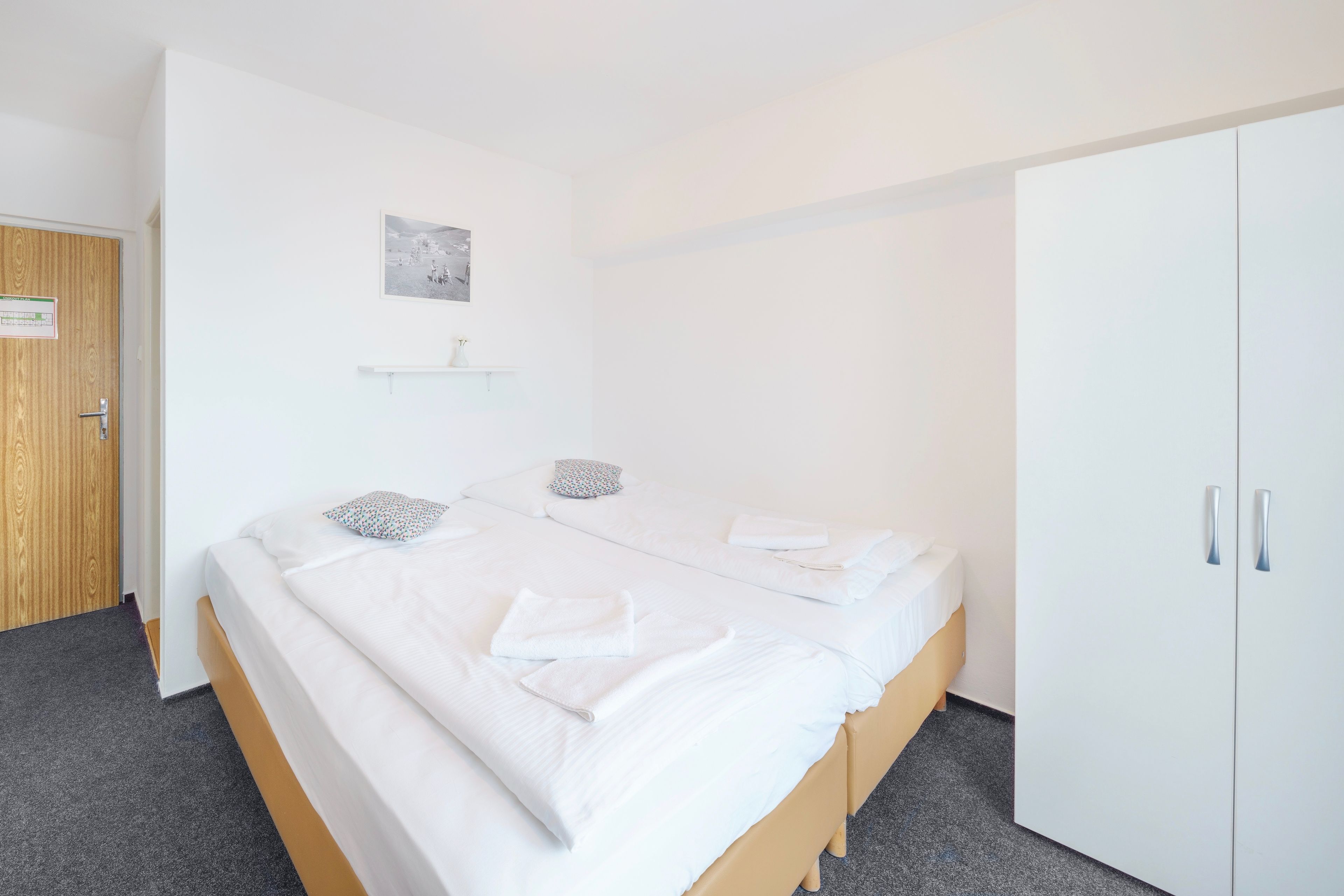 A bedroom with a bed, two pillows, a bedside table, a wardrobe, and a small shelf with a picture.