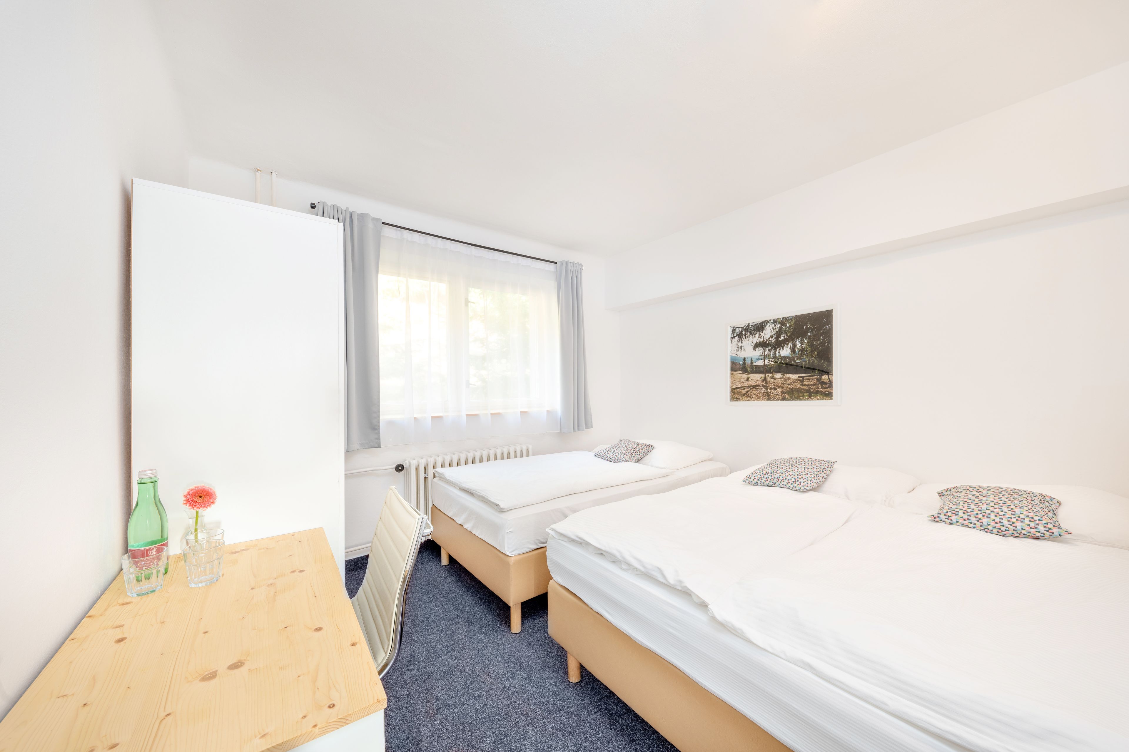 A hotel room with two single beds, a wooden table with a bottle and glass, a radiator, and a window with curtains.