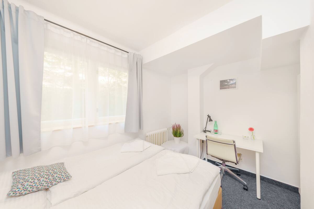 A hotel room with two beds, a desk, a radiator, and a window with curtains.