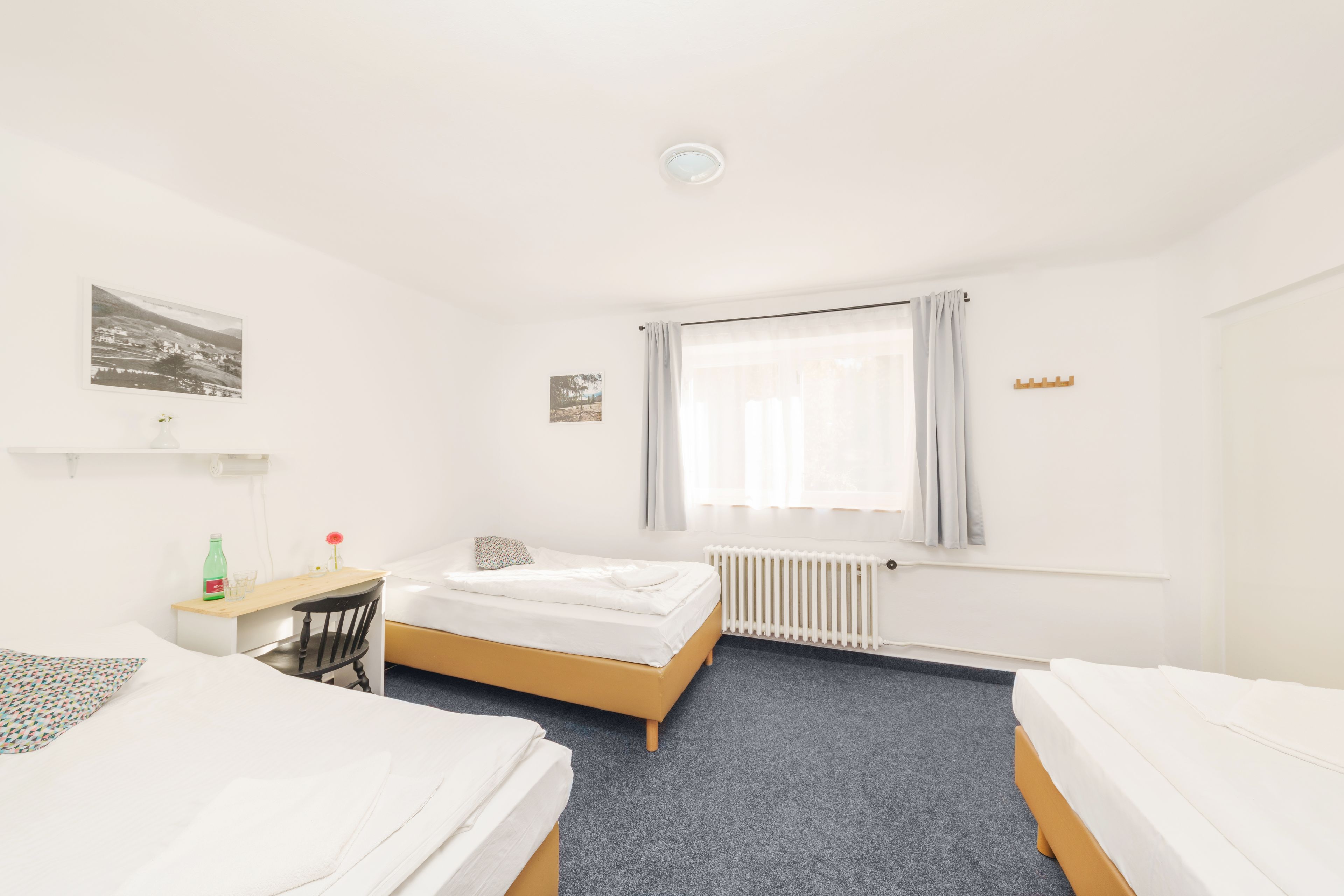 Habitació d'hotel amb dos llits, una taula de treball, una cadira, un radiador i una finestra amb cortines.
