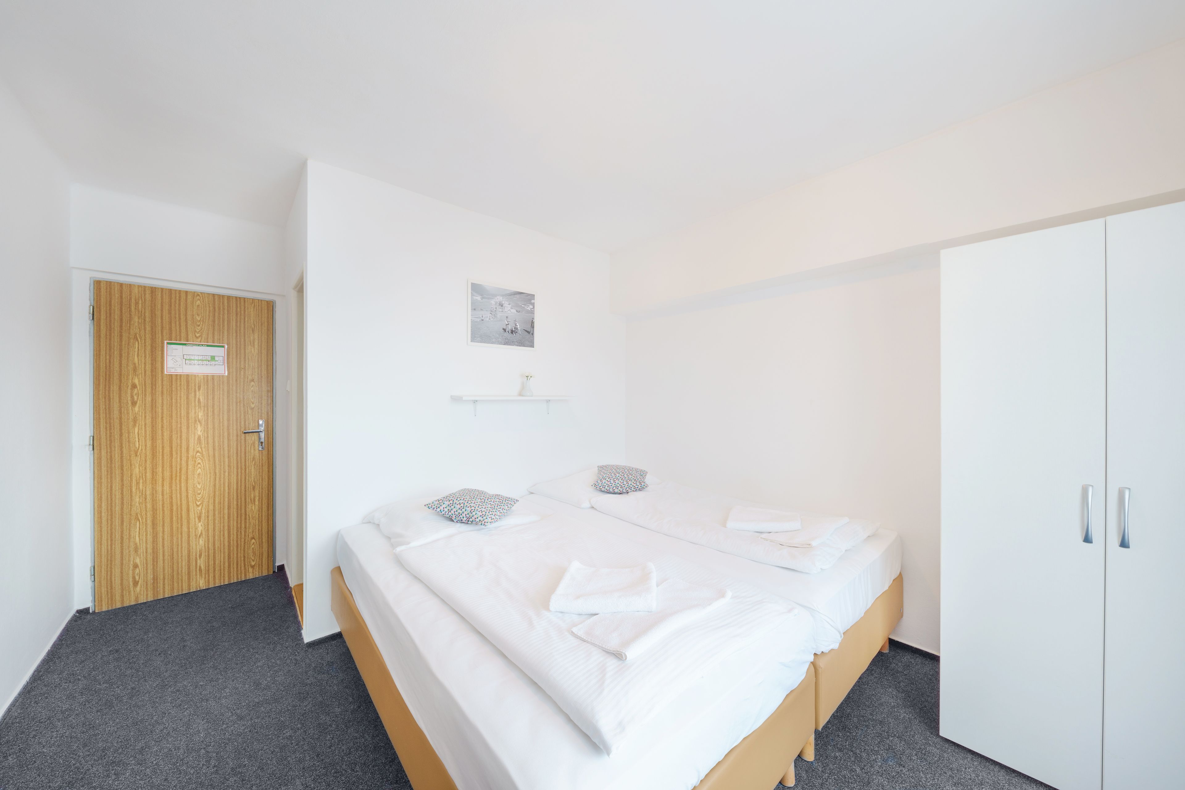 A hotel room with a wooden door, a bed with white linens, a small shelf with a picture, and a wardrobe.