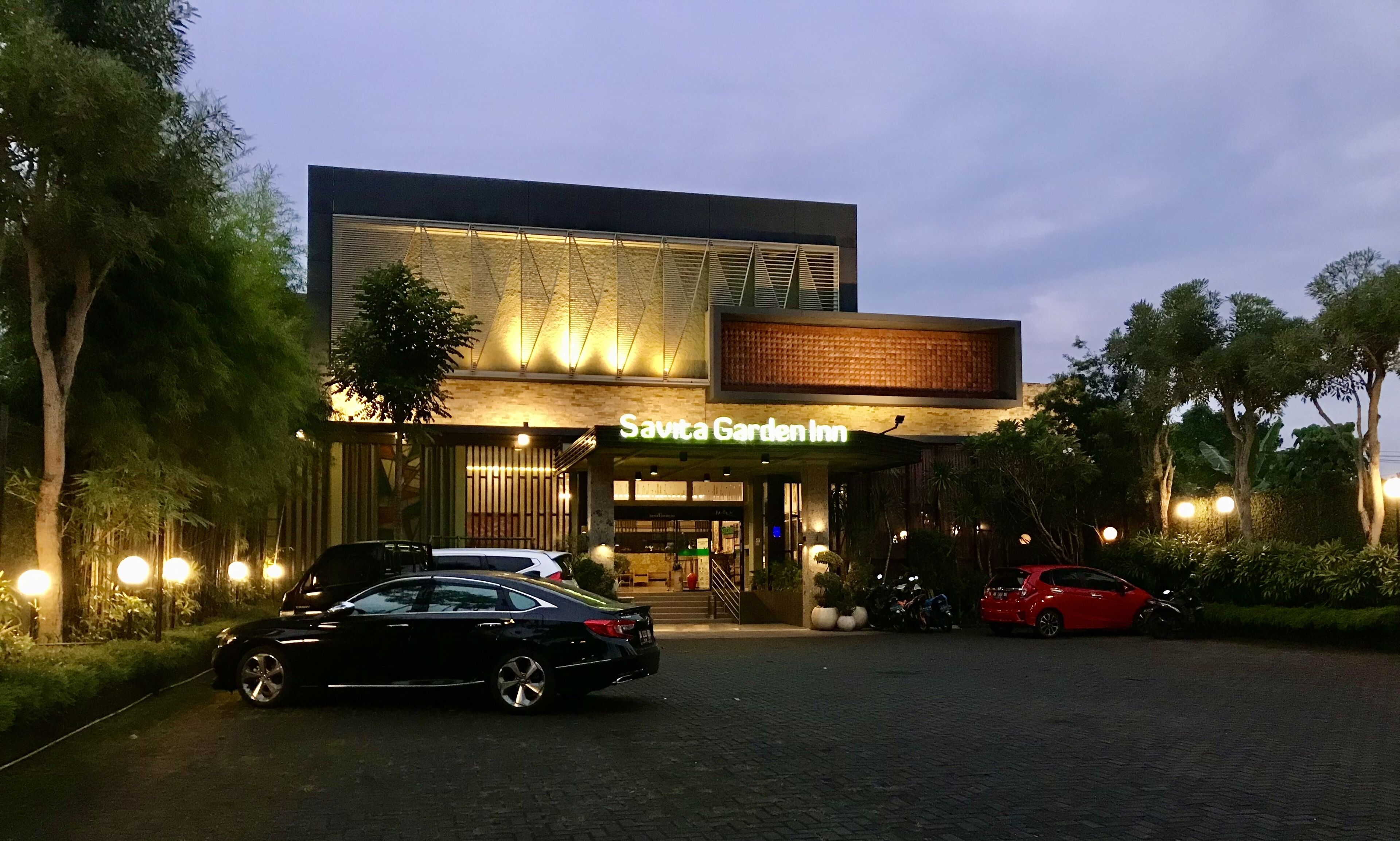 A modern building with a sign reading "Savita Garden Inn". There are cars parked in front and a well-lit entrance.