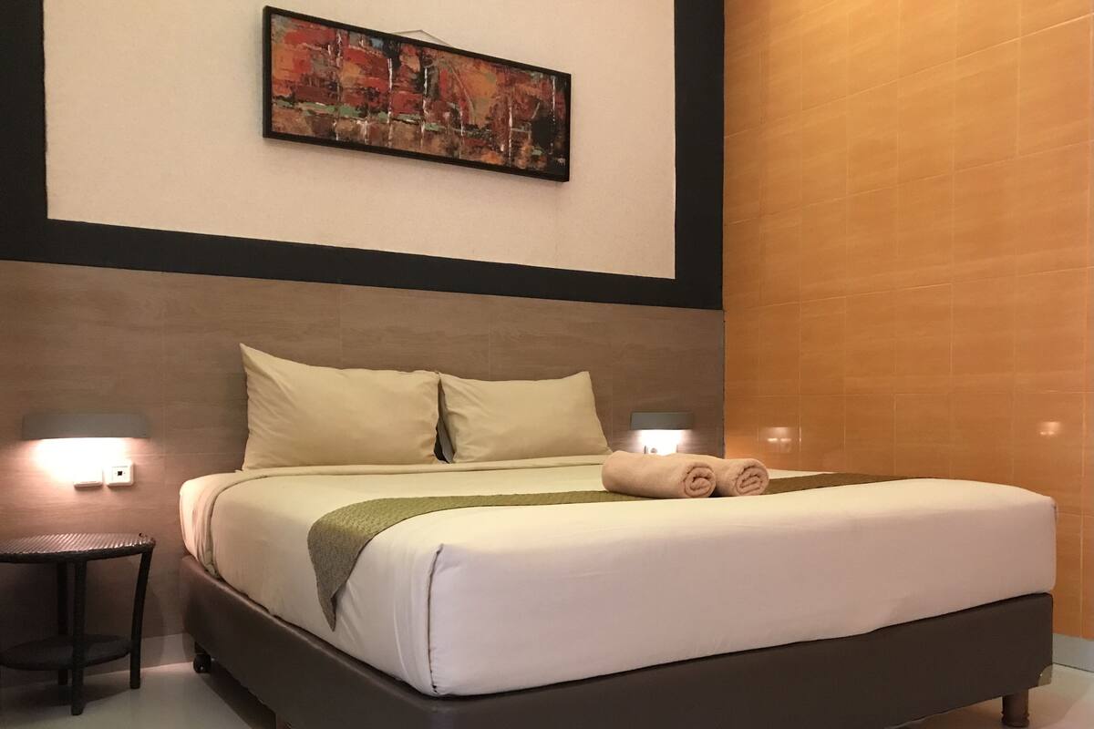 A neatly made bed with a white comforter and pillows, a green throw blanket, and a rolled towel on the side. There are two bedside tables with lamps, and a painting is visible on the wall above the bed.