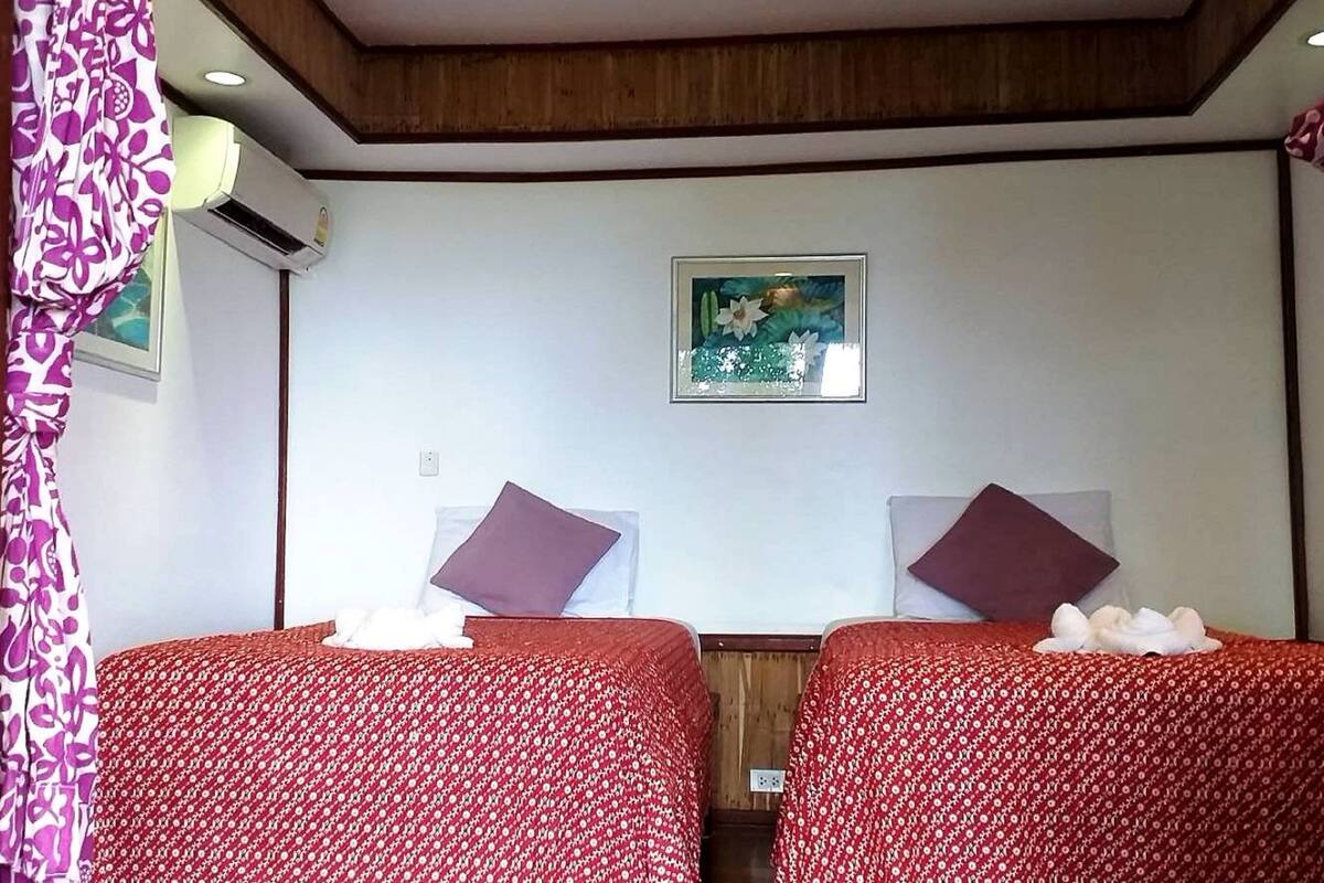 Une chambre d’hôtel avec deux lits, chacun recouvert d’une courtepointe à motifs floraux rouges, agrémenté d’oreillers violets et surmonté d’une petite table blanche avec une serviette. Un tableau encadré est accroché au mur, au-dessus des lits.