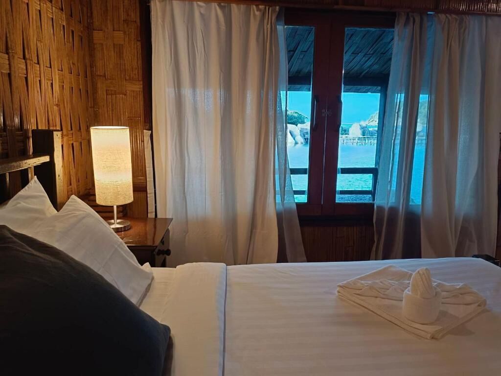 A hotel room with a bed, a nightstand with a lamp, and a view of the sea through large windows.