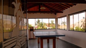 A spacious room with a ping pong table, large windows, and a view of palm trees. - Bougainville 1 Frente ao Mar de Peruíbe (Peruíbe)