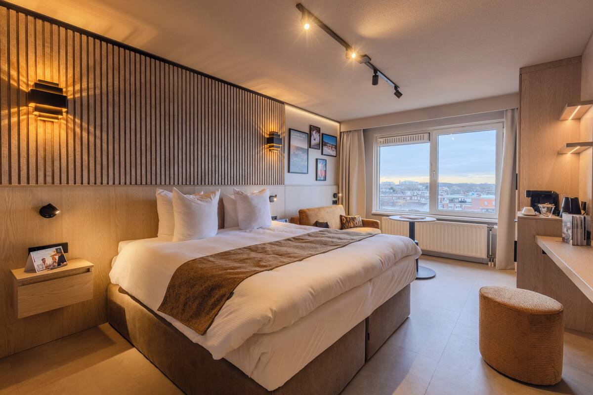 A modern hotel room with a large bed, bedside tables, a view of the city, and a small seating area.