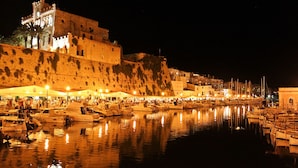 A historic waterfront area with a prominent castle-like building, a marina with boats, and a bustling street with outdoor seating. - My Rooms Ciutadella - Adults Only by My Rooms Hotels (Ciutadella de Menorca)