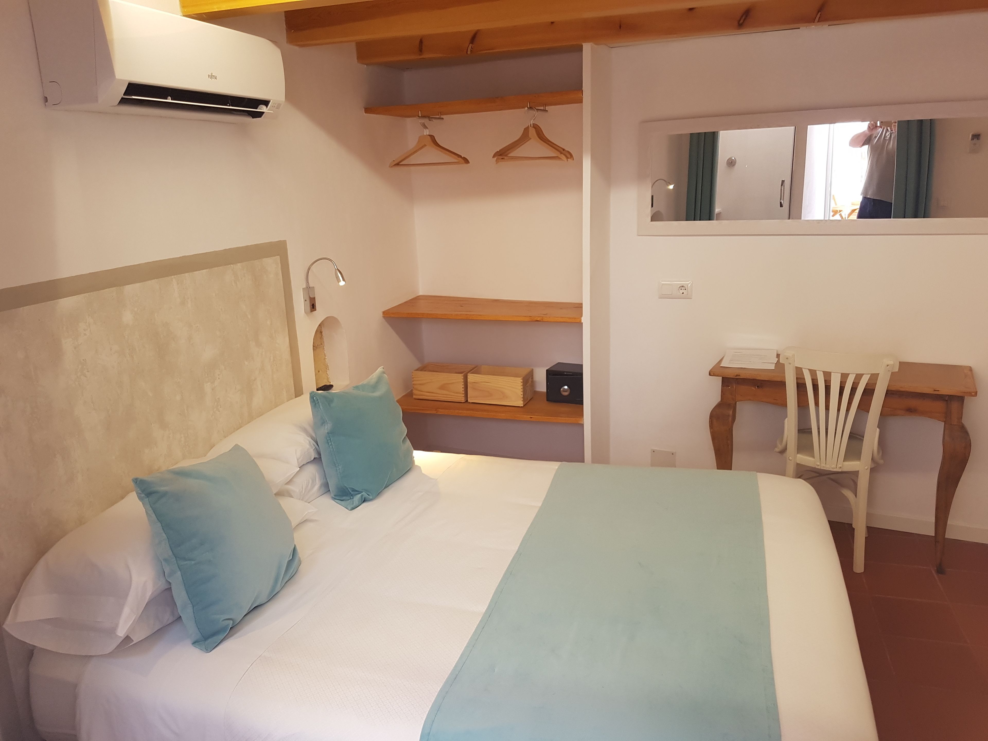 A bedroom with a bed, a desk, a chair, a mirror, and a shelf with hanging clothes.