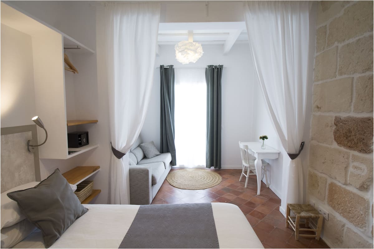 Ein Schlafzimmer mit einem Bett, einem Nachttisch, einem Sessel, einem kleinen Tisch mit Pflanze und Blick ins Freie durch Vorhänge.