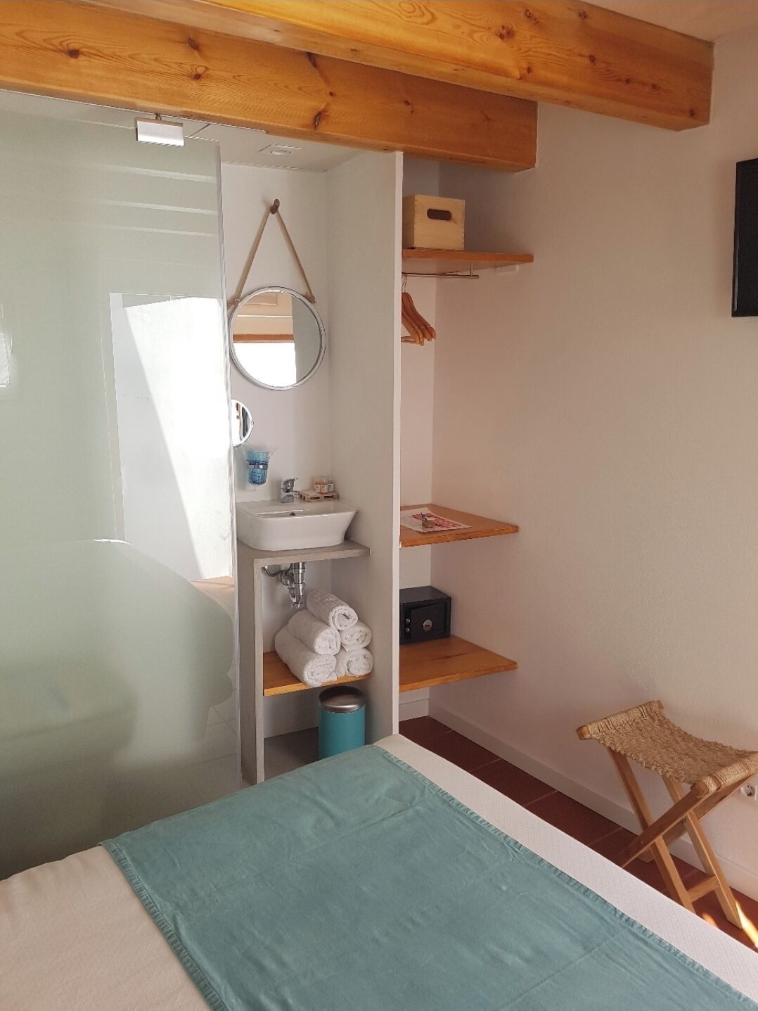 Una habitación pequeña y acogedora con una cama, una silla de madera, un lavamanos y estantes con toallas y artículos de aseo.