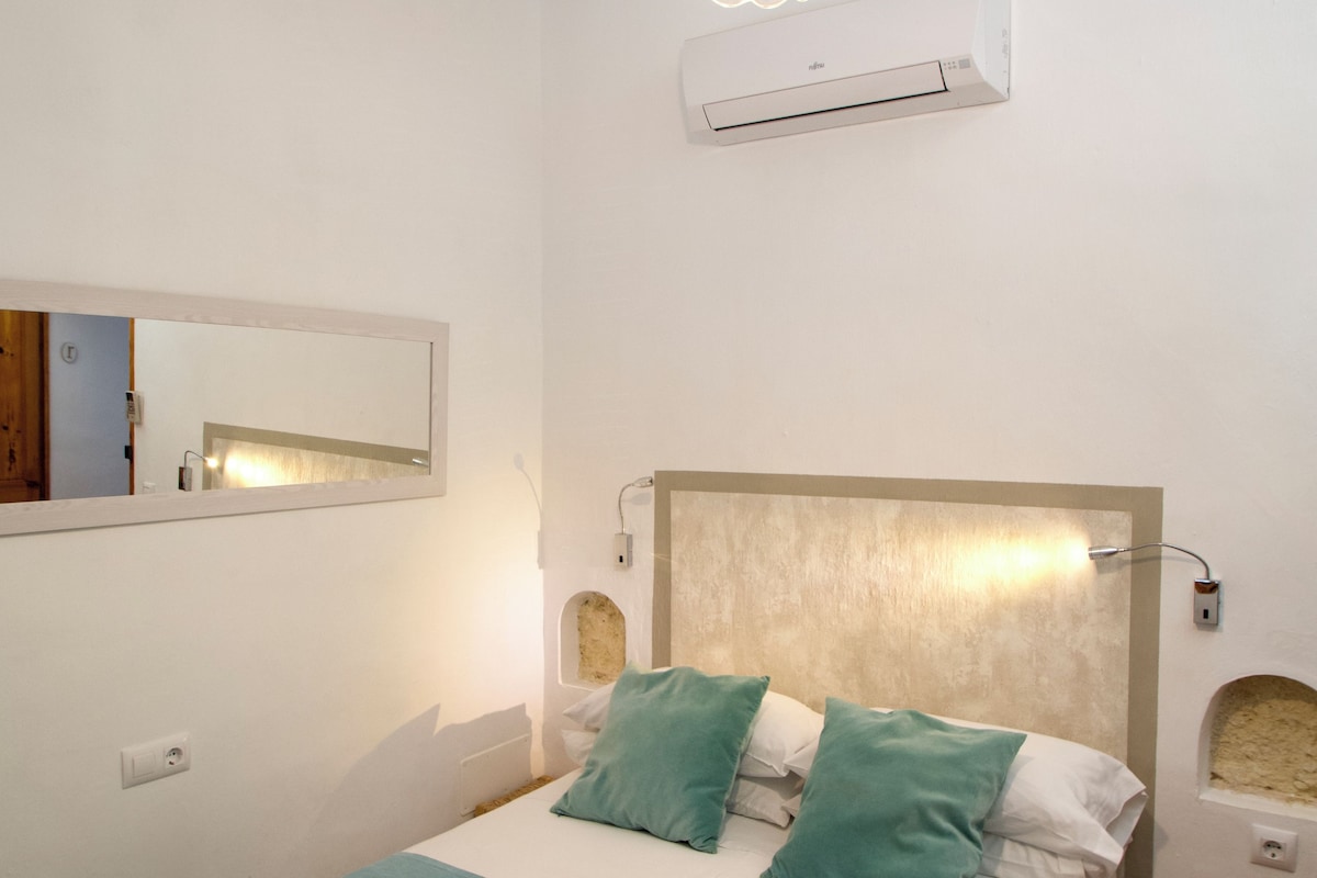 A bedroom with a bed, a bedside table, a mirror, and an air conditioning unit on the wall.