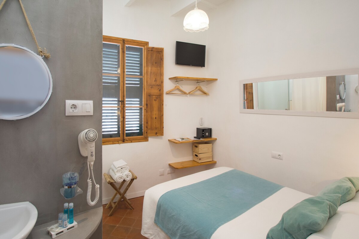 Une chambre à coucher comprenant un lit, une table de chevet, un luminaire suspendu, une fenêtre avec des stores, un téléviseur fixé au mur et un miroir.