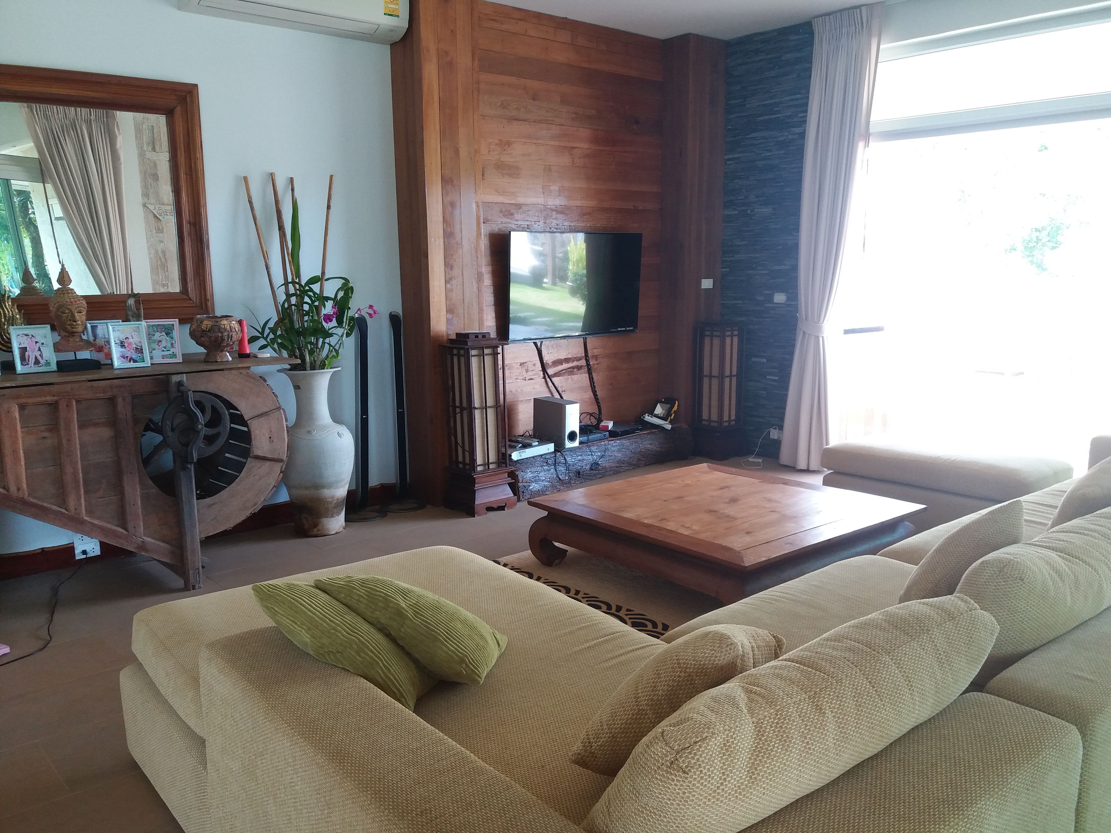 A living room with a sofa, a wooden coffee table, a TV mounted on a wooden wall, a large vase with flowers, and a cabinet with decorative items.