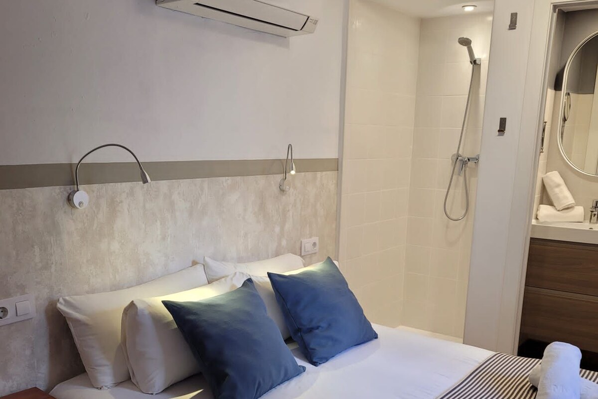 A hotel room with a bed, a bedside table, an air conditioning unit, a shower, and a mirror.