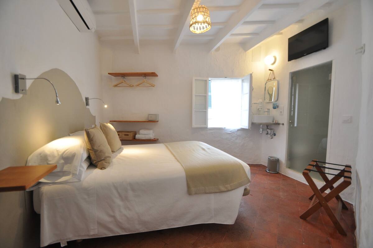 A bedroom with a bed, bedside table, hanging clothes rack, and a bathroom with a sink and mirror.