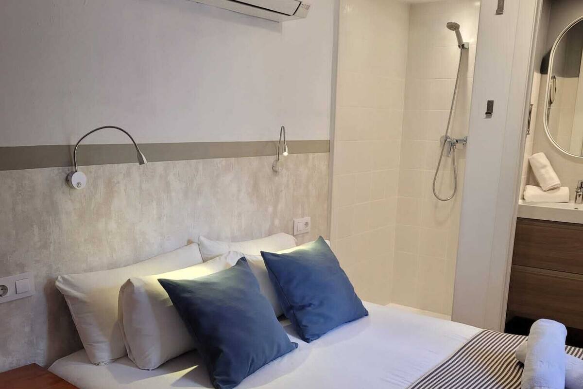 A hotel room with a bed, a bedside table, an air conditioning unit, a shower, and a mirror.