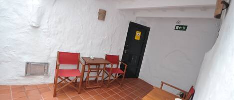 A small, cozy room with white walls and terracotta tiles. It includes a wooden table with chairs, a small window, and a door with an emergency exit sign.