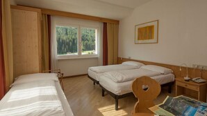 A room with two beds, a wooden headboard, a desk with a lamp, and a window with a view of trees.