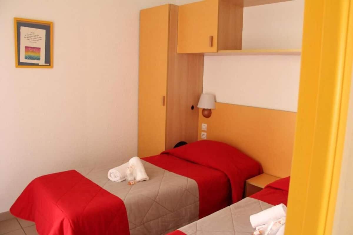 Habitación de hotel con dos camas, cada una con colcha roja y toalla blanca. Encima de las camas hay un armario de madera y una mesita de noche con lámpara.