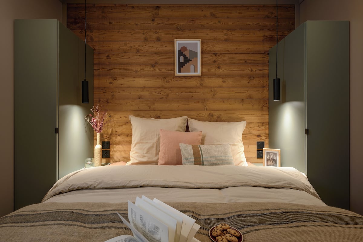 Un dormitorio moderno con una pared de madera, una cama con muchas almohadas, una mesita de noche con un jarrón y un marco de fotos, y un tazón de galletas sobre la cama.