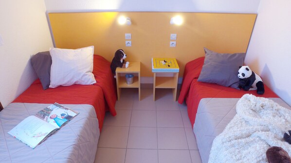 Ein Zimmer mit zwei Betten, jedes mit einem Kopfkissen und einer Decke. Ein kleiner Tisch mit einem Spielzeug und einem Buch.
