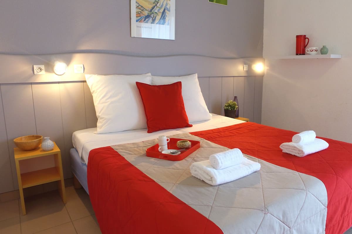 Una cama bien hecha con ropa de cama roja y blanca, una bandeja con tetera y taza, y una pequeña mesita de noche con un cuenco y un florero.