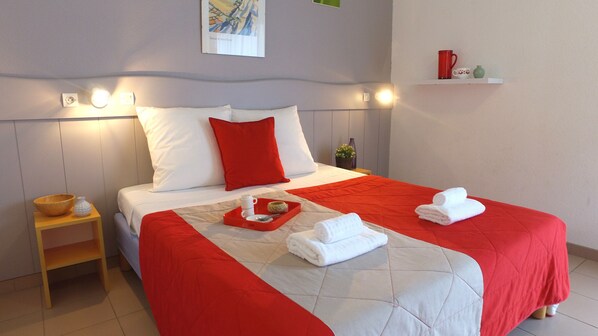 A neatly made bed with red and white bedding, a tray with a teapot and cup, and a small wooden nightstand with a bowl and vase.