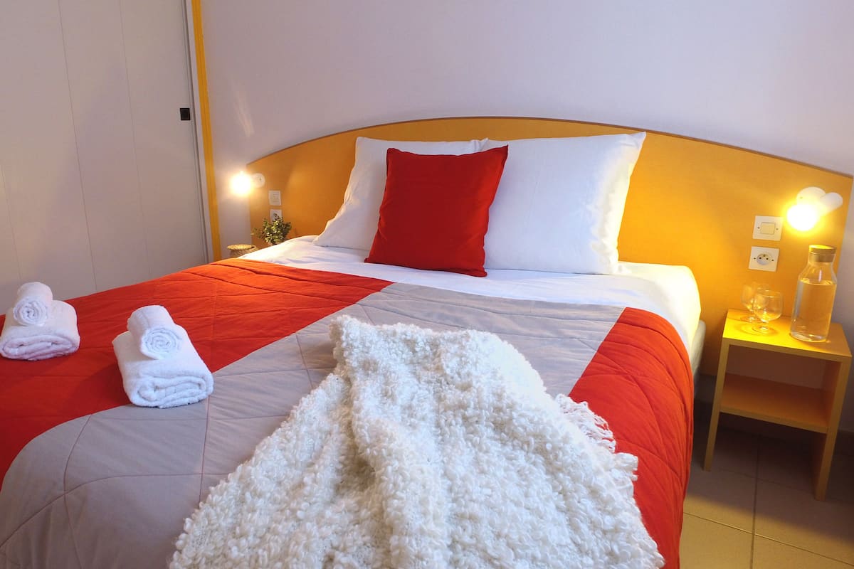 Habitación de hotel con una cama, dos toallas enrolladas, un edredón blanco y esponjoso, y una mesita de noche con una botella y vasos.