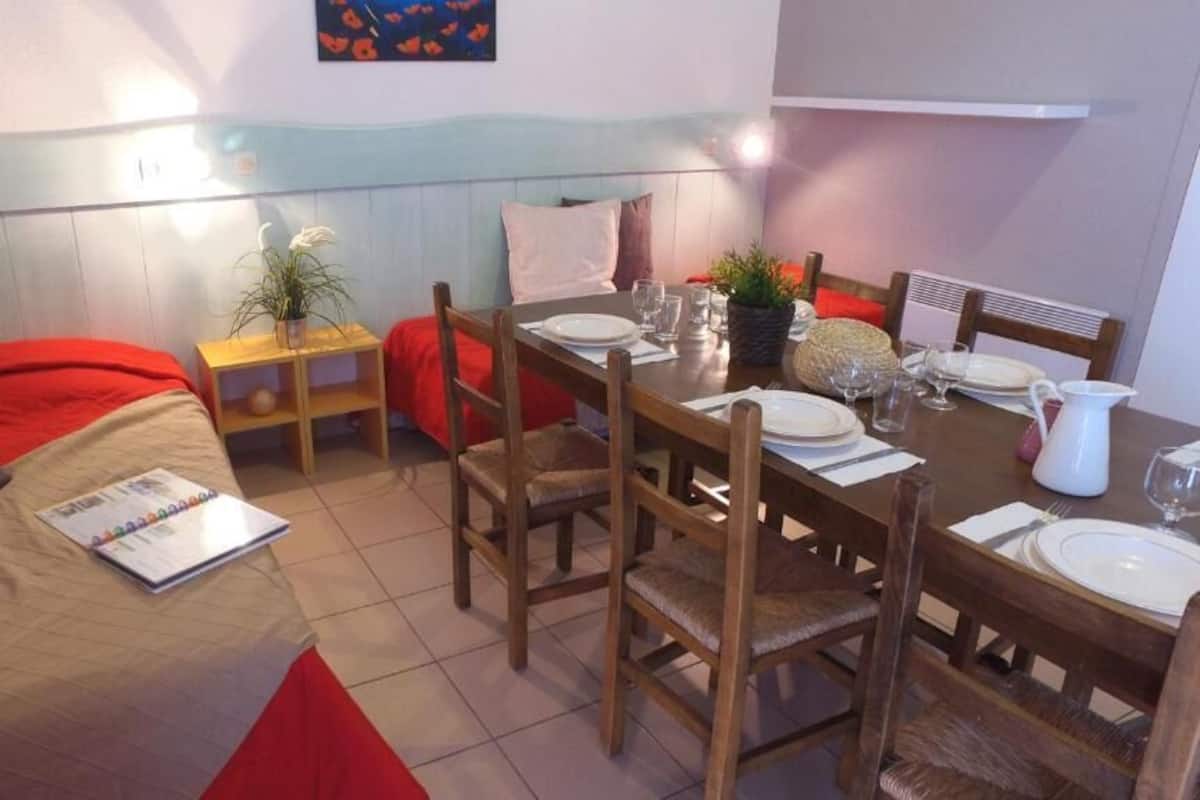 Una habitación compacta con una cama, una mesa de comedor para dos personas y una mesita auxiliar con una planta.