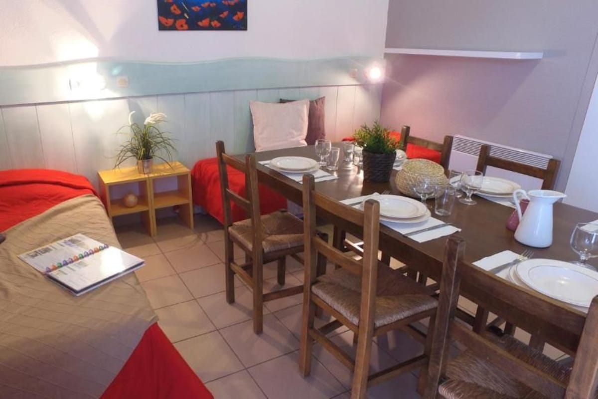 Une pièce de taille modeste comprenant un lit, une table à manger dressée pour deux personnes et une petite table d’appoint agrémentée d’une plante.