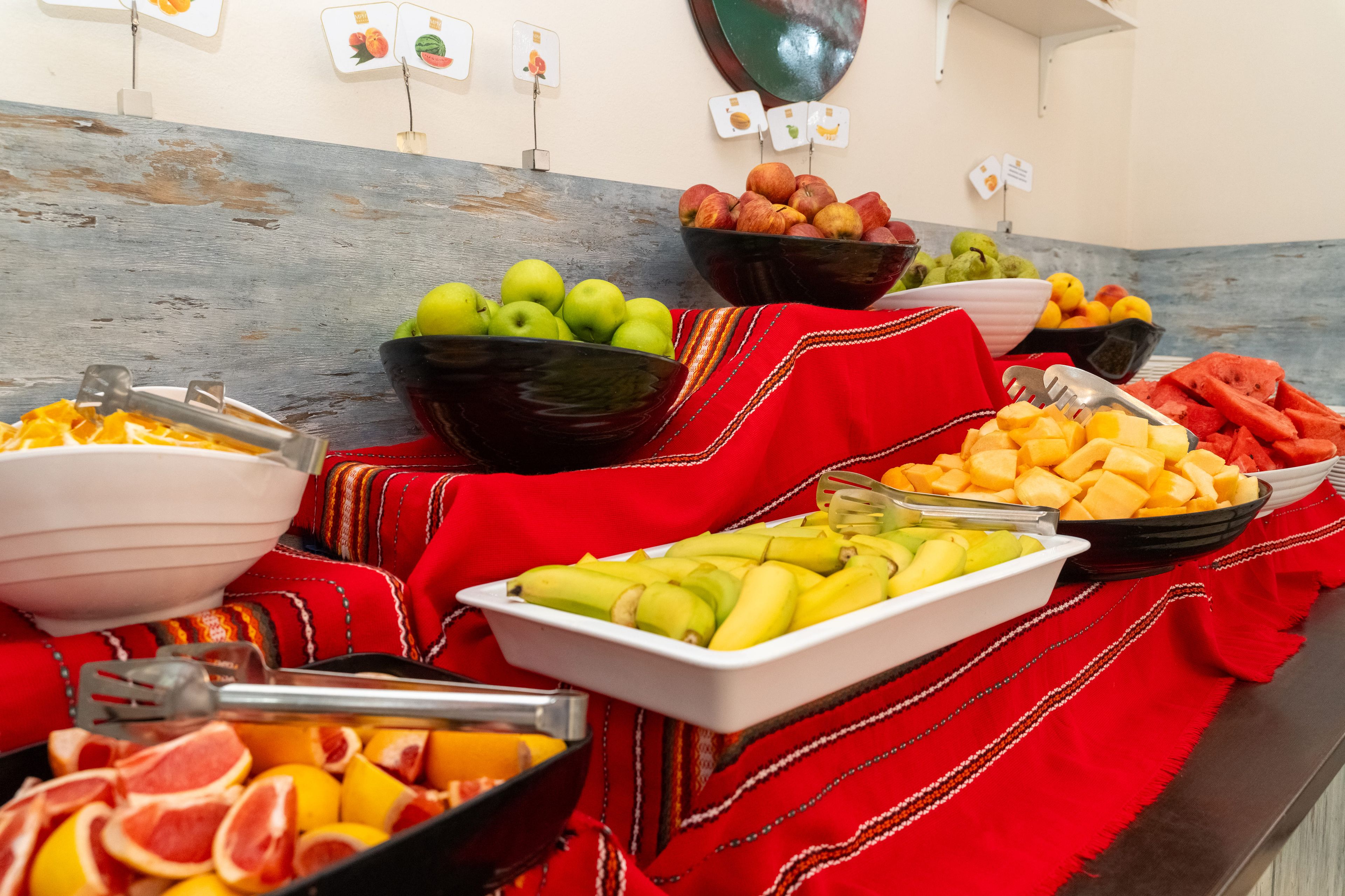Una mesa de buffet con diversas frutas y verduras, incluyendo manzanas, plátanos y sandía, dispuestas sobre un mantel rojo.